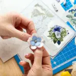 Hands working on a lovely scrapbook, including a digital print of a beautiful blue flower. Visit our shop at DigitalFlowersCrafts.etsy.com